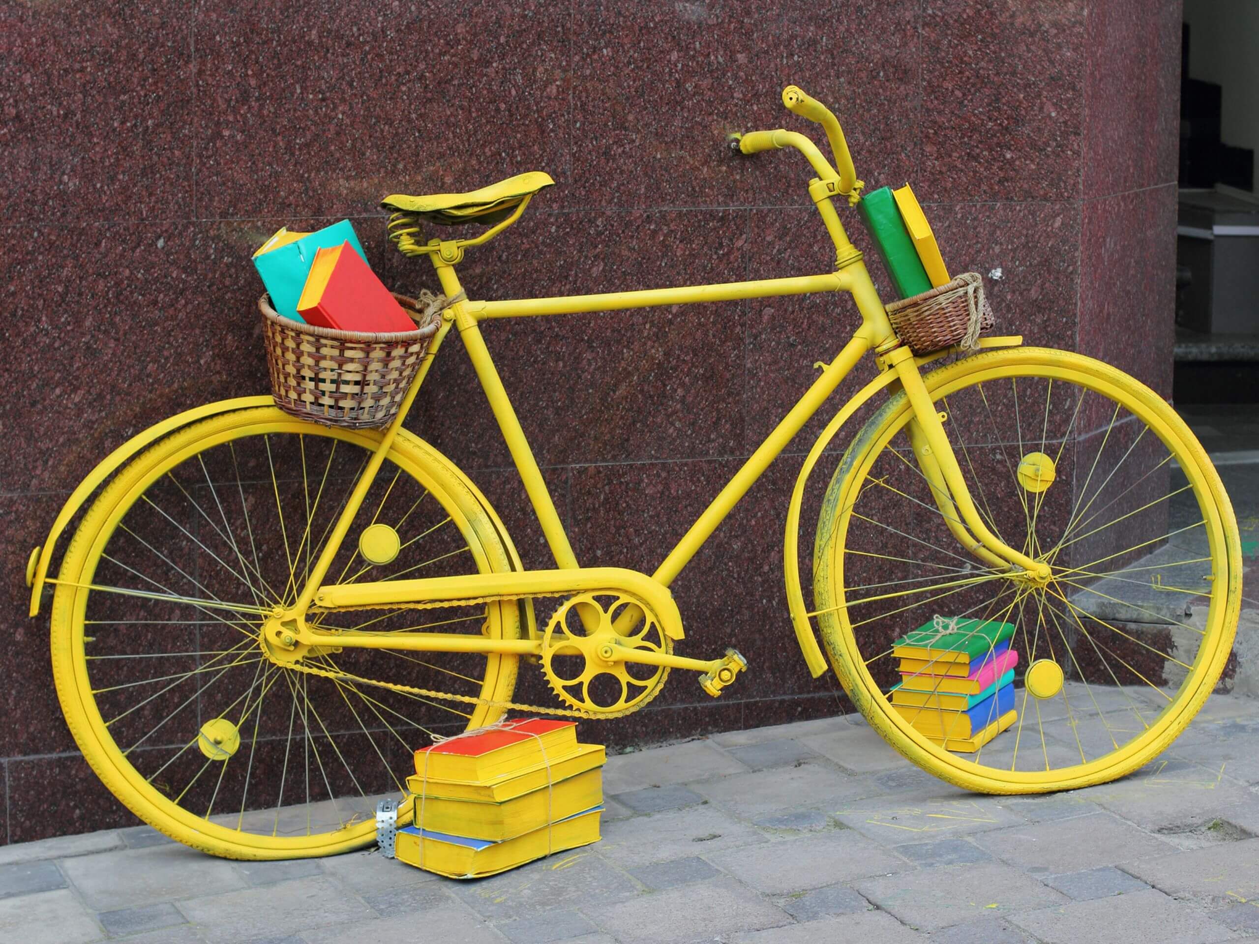 bicycle leans against brick wall. The entire bike is painted bright yellos, including a pile of books sitting beside it.